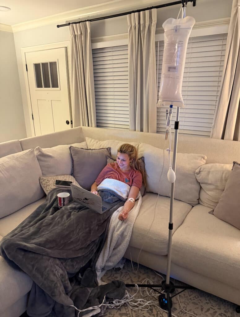 Woman receiving IV therapy while using a laptop on a couch, surrounded by blankets, highlighting hydration and wellness at home.