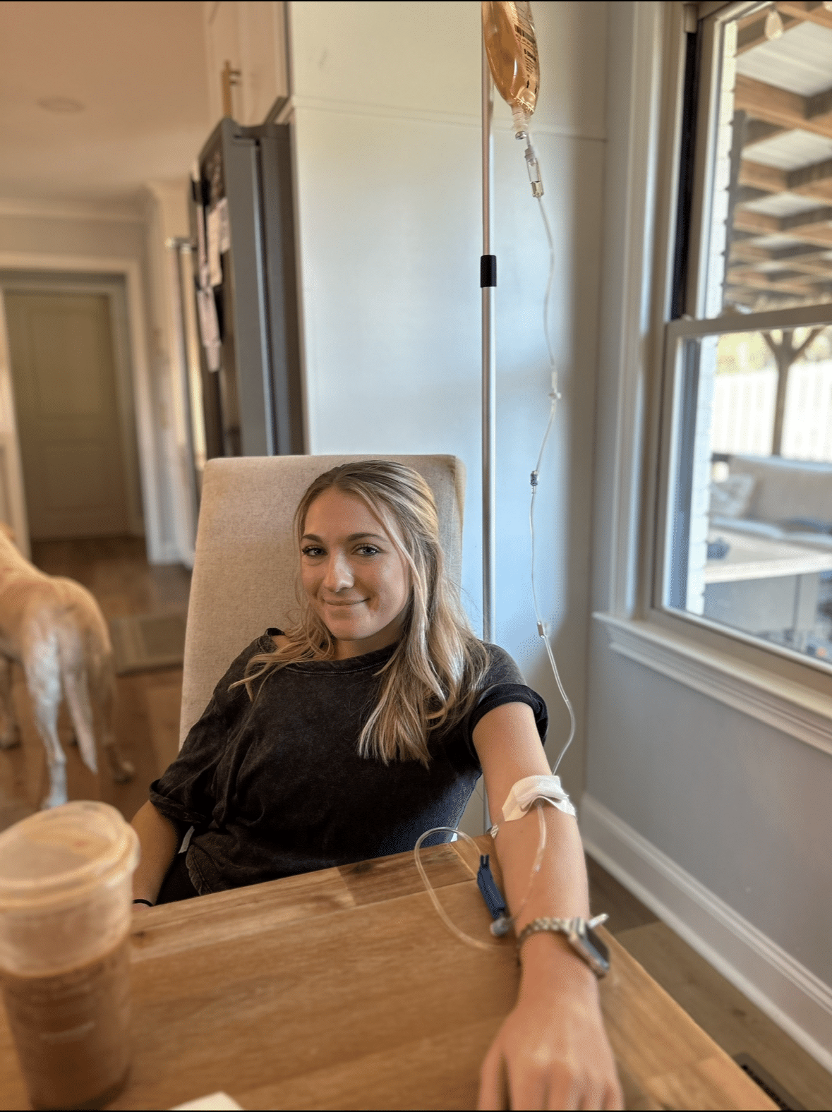 Woman receiving mobile IV therapy at home, smiling, with IV drip in arm, casual setting with a drink on table.