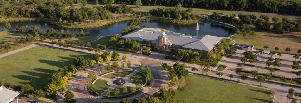 Aerial view of Keller Town Hall in Keller, Texas, surrounded by green lawns, walking paths, a circular plaza, and a peaceful pond with a fountain, set against a backdrop of dense trees